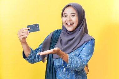 Sarı arka planda tesettürlü gülümseyen kadın kredi kartını tutuyor..