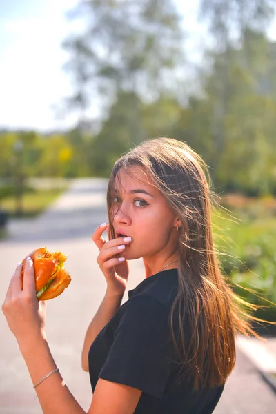 Woman eating burger and fries smiling. Beautiful caucasian female model eating a hamburger with hands over blurred background outside. Still life, eating out concept.