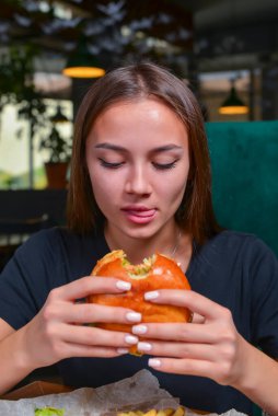 Hamburger yiyen ve patates kızartması yiyen bir kadın. Kafkasyalı güzel bir kadın model elleri bulanık bir restoranın arka planında hamburger yiyor. Hayat, dışarıda yemek kavramı..