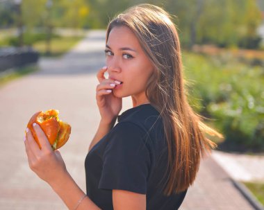 Hamburger yiyen ve patates kızartması yiyen bir kadın. Güzel beyaz kadın manken elleri dışarıda bulanık bir şekilde hamburger yiyor. Hayat, dışarıda yemek kavramı..