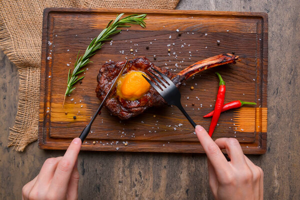 Barbecue tomahawk steak with spices served on a rustic wooden board over wooden background. BBQ concept, grilled meat steak, autumn seasonal dish. Eating steak with knife and fork.