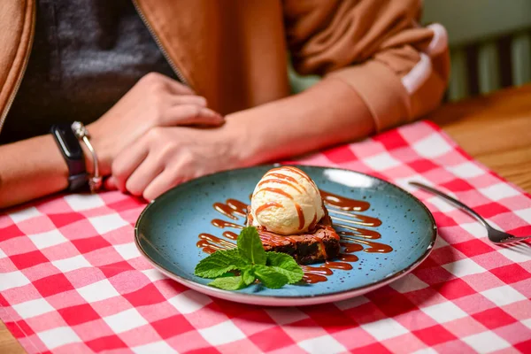 Traditional recipe brownie with ice cream served on a blue plate over table with red plaid tablecloth. Italian cuisine concept, sweet dessert. Young woman eating chocolate brownie and ice cream.