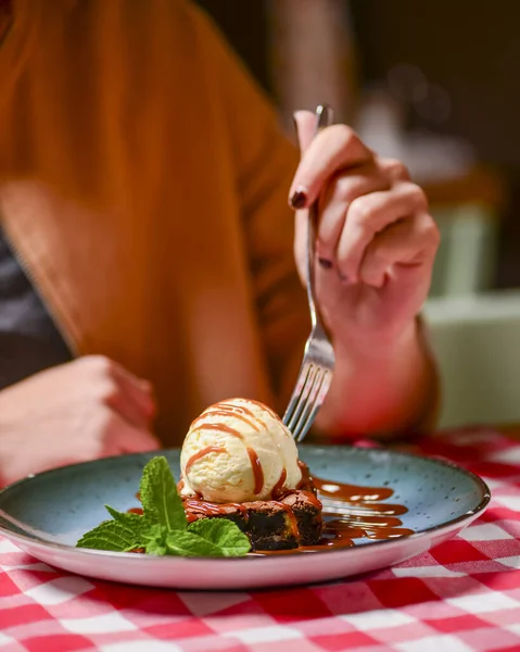 Traditional recipe brownie with ice cream served on a blue plate over table with red plaid tablecloth. Italian cuisine concept, sweet dessert. Young woman eating chocolate brownie and ice cream.