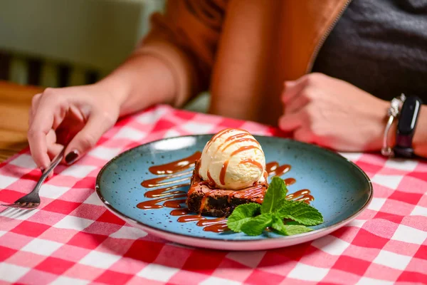 Traditional recipe brownie with ice cream served on a blue plate over table with red plaid tablecloth. Italian cuisine concept, sweet dessert. Young woman eating chocolate brownie and ice cream.