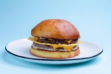 Sade pastel mavi arka planda beyaz bir tabakta servis edilen jambonlu ve peynirli dev mükemmel hamburger. Kapatın. Fast food, abur cubur kavramı.
