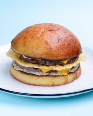 Sade pastel mavi arka planda beyaz bir tabakta servis edilen jambonlu ve peynirli dev mükemmel hamburger. Kapatın. Fast food, abur cubur kavramı.