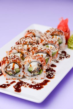 Yılanbalığı ve unagi soslu Japon suşisi seti. İçinde krem peynir var. Pastel renkli mor arka planda beyaz bir tabakta servis edilmiş. Geleneksel tarif.