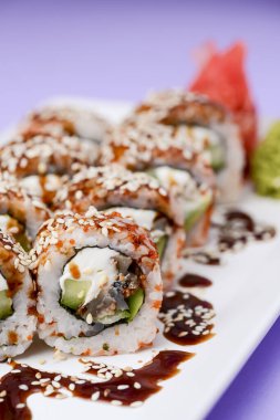 Yılanbalığı ve unagi soslu Japon suşisi seti. İçinde krem peynir var. Pastel renkli mor arka planda beyaz bir tabakta servis edilmiş. Geleneksel tarif.