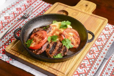 Geleneksel Ukrayna masa örtüsünün üzerinde ızgara domatesli bir tavada servis edilen sulu biftek. Ukrayna mutfağı kavramı, barbekü kavramı.