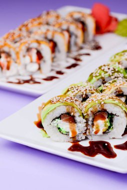 Maki suşi rulosu füme yılan balığı, krem peynir ve unagi sosundan yapılır. Geleneksel Japon mutfağı kavramı. Mor arka planda beyaz bir tabakta servis edilmiş..