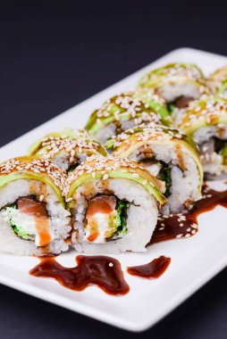 Uramaki suşi rulosu tütsülenmiş yılan balığı ve avokado ile kaplanmış unagi ve siyah arka planda beyaz tabakta susam tohumları. Japon mutfağı kavramı.