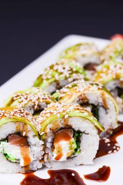 Uramaki suşi rulosu tütsülenmiş yılan balığı ve avokado ile kaplanmış unagi ve siyah arka planda beyaz tabakta susam tohumları. Japon mutfağı kavramı.