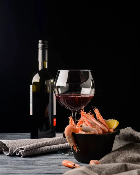 Shrimps with lemon and fresh dill in a small black bowl and a bottle of red wine over black background. Sea food, healthy cuisine concept. Close up.