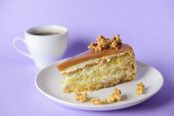 One piece of Caramel cake with pop corn on top served on a white plate. Over pastel violet purple background. Focus on a piece of cake.