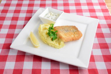 Kızarmış balık ve patates püresi kırmızı ekose masa örtüsünün üzerinde beyaz kare bir tabakta servis ediliyor. İtalyan mutfağı, akşam yemeği fikri..