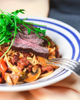 Yumurtalı makarna, sığır eti, mantar ve taze biberiye ve roka ile süslenmiş kremalı makarna. Masa örtüsüyle ahşap arka planda beyaz bir tabakta servis edilmiş. İtalyan mutfağı kavramı.