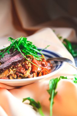 Yumurtalı makarna, sığır eti, mantar ve taze biberiye ve roka ile süslenmiş kremalı makarna. Masa örtüsüyle ahşap arka planda beyaz bir tabakta servis edilmiş. İtalyan mutfağı kavramı.