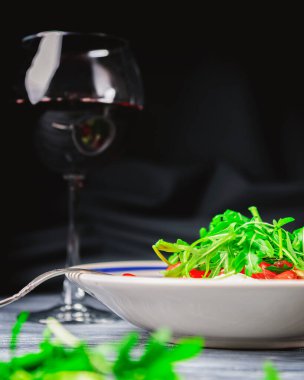 Domatesli salata ve üstüne taze roka ve bir bardak kırmızı şarap. İtalyan mutfağı konsepti. Beyaz tabakta servis edilen salata gri ahşap arka planda ve siyah masa örtüsüyle servis ediliyor..