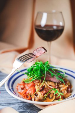 Yumurtalı erişte, biftek, mantar ve taze biberiye, roka ve bir bardak kırmızı şarapla süslenmiş kremalı biftek makarnası. Ahşap arka planda masa örtüsüyle beyaz bir tabakta servis edilmiş..