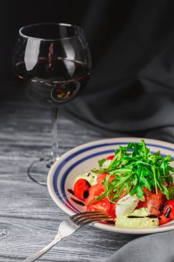 Domatesli salata ve üstüne taze roka ve bir bardak kırmızı şarap. İtalyan mutfağı konsepti. Beyaz tabakta servis edilen salata gri ahşap arka planda ve siyah masa örtüsüyle servis ediliyor..