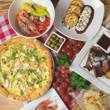 Geleneksel İtalyan mutfağı konsepti düzdü. Deniz ürünleri pizza, pişmiş karides, karamelli pirzola, jambonlu sandviç, yeşil zeytin ve kırmızı ekose masa örtüsünün üzerinde taze vişneli domates..