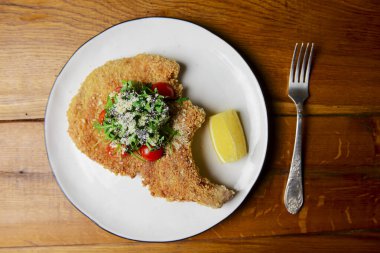 Üzerinde kiraz domates ve taze roka olan bol yağda kızartılmış balık kemiği bifteği. Kırsal ahşap arka planda beyaz bir tabakta bir parça limonla servis edilmiş..