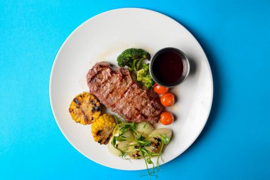 Lezzetli ızgara biftek, bol sebzeli ve mısır, beyaz bir tabakta servis ediliyor ve mavi arka planda sos var. BBQ konsepti.