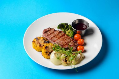 Lezzetli ızgara biftek, bol sebzeli ve mısır, beyaz bir tabakta servis ediliyor ve mavi arka planda sos var. BBQ konsepti.