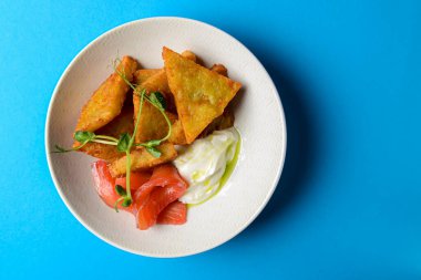 Somon balığı ve ekşi krema sosuyla servis edilen taze kızarmış patatesli krep. Mavi arka planda beyaz bir kasede.