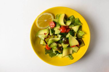 Salatalıktan yapılmış sebze salatası, kiraz domatesleri, avokado, marul salatası ve limon beyaz arka planda parlak sarı bir kasede servis edilir. Sağlıklı yeme kavramı.