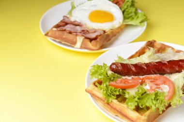 Waffle sağlıklı kahvaltı. Izgara sosisli ekşi waffle, kızarmış yumurta, lezzetli sos ve taze sebze. Waffle üzerinde marul salatası olan sebzeler. Parlak sarı arkaplanda servis edilir.