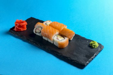 Philadelphia Maki Suşi taze çiğ somon, krem peynir ve salatalıktan yapılmıştır. Mavi arka planda siyah tahtada servis edilir. Japon mutfağı kavramı.