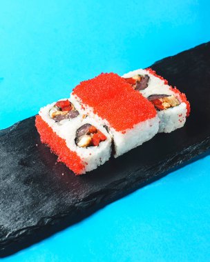 Tobiko Baharatlı Maki Suşi - Tobiko dışarıda, somon içeride. Kırmızı havyar içinde yuvarlanıyor, siyah bir tabakta, mavi arka planda, izole edilmiş..