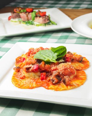 Beyaz tabakta ıspanaklı ve peynirli İtalyan mantısı. Yeşil ekose masa örtüsüyle taşralı ahşap bir masada servis edilmiş. İtalyan mutfağı konsepti, köy tarzı..