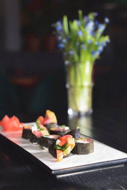 Maki suşisi siyah arka planda beyaz bir tabak üzerine kurulmuş. Japon mutfağı konsepti, zencefil ve wasabi soslu maki dürümleri. Arka planda bahar çiçekleri.