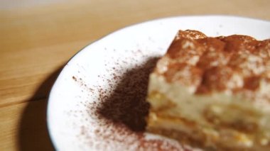 Kamera tiramisu kekinin üzerinde yavaşça hareket ediyor, tiramisu kekine odaklanıyor. Ağır çekim. Beyaz tabakta, ahşap masada tatlı İtalyan tatlısı. Çok yakın çekim..