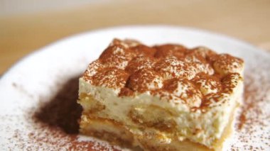 Kamera tiramisu kekinin üzerinde yavaşça hareket ediyor, tiramisu kekine odaklanıyor. Ağır çekim. Beyaz tabakta, ahşap masada tatlı İtalyan tatlısı. Çok yakın çekim..