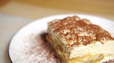 Kamera tiramisu kekinin üzerinde yavaşça hareket ediyor, tiramisu kekine odaklanıyor. Ağır çekim. Beyaz tabakta, ahşap masada tatlı İtalyan tatlısı. Çok yakın çekim..