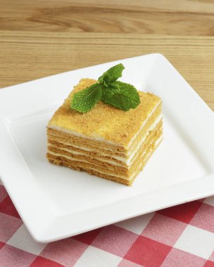 Üstünde nane yaprağı olan bir tabak ballı kek. Kırmızı ekose arka planda beyaz bir tabakta servis edilmiş. Çay ya da kahve için bal tatlısı..
