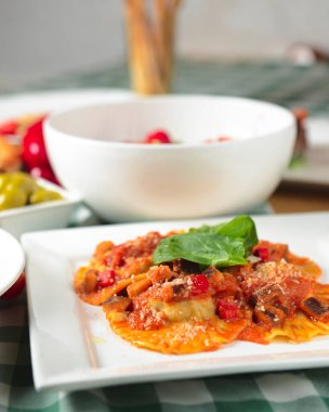 Beyaz tabakta ıspanaklı ve peynirli İtalyan mantısı. Yeşil ekose masa örtüsüyle taşralı ahşap bir masada servis edilmiş. İtalyan mutfağı konsepti, köy tarzı..