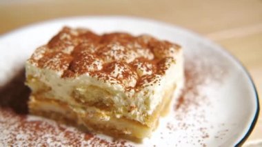 Kamera tiramisu kekinin üzerinde yavaşça hareket ediyor, tiramisu kekine odaklanıyor. Ağır çekim. Beyaz tabakta, ahşap masada tatlı İtalyan tatlısı. Çok yakın çekim..