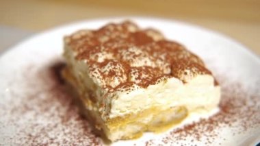 Kamera tiramisu kekinin üzerinde yavaşça hareket ediyor, tiramisu kekine odaklanıyor. Ağır çekim. Beyaz tabakta, ahşap masada tatlı İtalyan tatlısı. Çok yakın çekim..