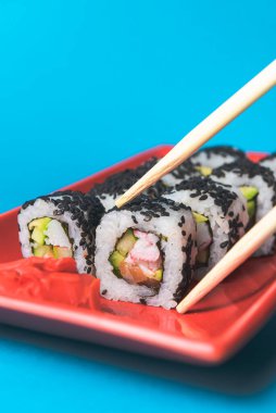 Lezzetli ve taze suşi seti ahşap çubuklarla kırmızı bir tabakta servis ediliyor. Açık mavi arkaplanda. Yakın plan, suşi üzerine seçici odaklanma. Kaliforniya ya da Philadelphia suşi seti.