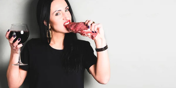 Beautiful aggressive woman bites raw meat with her teeth. Caucasian woman with black hair biting raw meat and drinking red wine. Creative art concept.