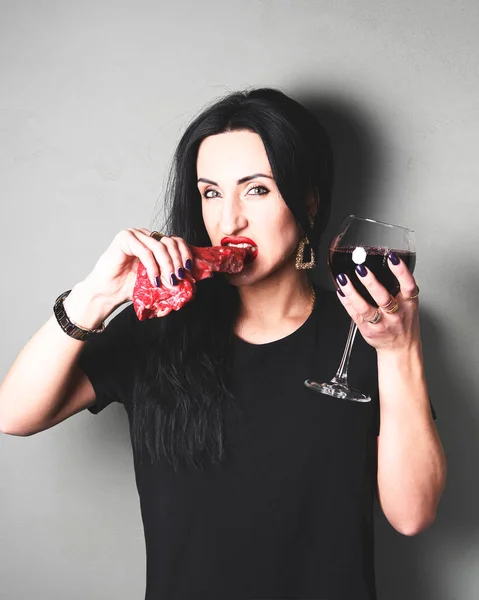 Beautiful aggressive woman bites raw meat with her teeth. Caucasian woman with black hair biting raw meat and drinking red wine. Creative art concept.