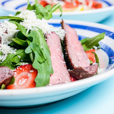 Beyaz bir tabakta servis edilen sığır eti, çilek, domates, taze roka ve peynirli salata. Pastel mavi arka planda. Sağlıklı yeme kavramı.