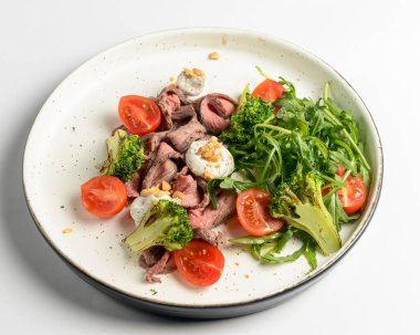 Beyaz arka planda servis edilen et, kiraz domatesleri, brokoli, keçi peyniri ve taze roka salatasından yapılmış sağlıklı bir salata. Lezzetli öğle yemeği ya da akşam yemeği fikri.