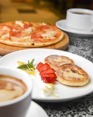 Naneli, çilekli ve ekşi kremalı peynirli kek. Beyaz tabakta bir fincan kahveyle servis edilen tatlı. Sebze ve tavuklu pizza da masada, iki fincan çay..