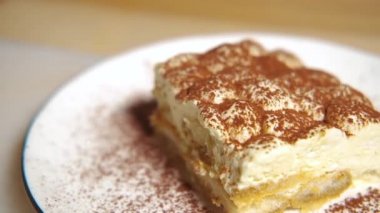 Kamera tiramisu kekinin üzerinde yavaşça hareket ediyor, tiramisu kekine odaklanıyor. Ağır çekim. Beyaz bir tabakta, ahşap bir masada tatlı geleneksel İtalyan tatlısı..