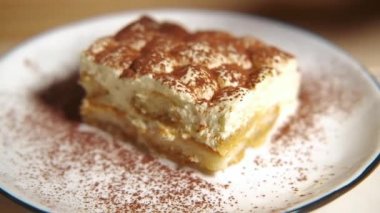 Kamera tiramisu kekinin üzerinde yavaşça hareket ediyor, tiramisu kekine odaklanıyor. Ağır çekim. Beyaz bir tabakta, ahşap bir masada tatlı geleneksel İtalyan tatlısı..